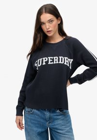 Marineblaues Sweatshirt mit weißem "SUPERDRY"-Text, gerippter Textur, Rundhalsausschnitt und langen Raglanärmeln. Kombiniert mit hellblauen Jeans.