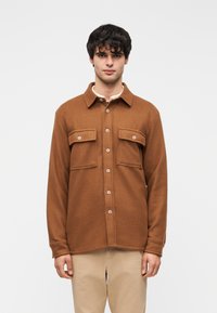 Καφέ πουκάμισο από φανέλα με δύο τσέπες στο στήθος και κλείσιμο με κουμπιά. Απαλή υφή, κλασικό γιακά, μακριά μανίκια και ίσια γραμμή στο τελείωμα.