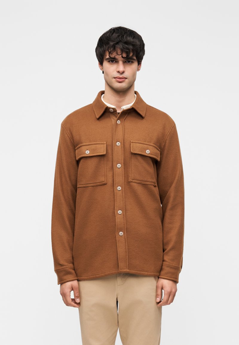 Καφέ πουκάμισο από φανέλα με δύο τσέπες στο στήθος και κλείσιμο με κουμπιά. Απαλή υφή, κλασικό γιακά, μακριά μανίκια και ίσια γραμμή στο τελείωμα.