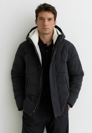 Czarna pikowana kurtka puchowa z dużym kapturem wyłożonym kremowym polarem oraz przednim zamkiem błyskawicznym. Rękawy o prostym kroju z mankietami.