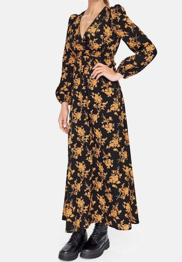 Kvinna klädd i en svart långärmad blommig maxiklänning med orange-gula blommor, kombinerat med svarta snörstövlar.