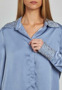 Blusa de satén azul claro con cuello y puños adornados con tachuelas plateadas, que presenta una textura suave y un corte entallado.