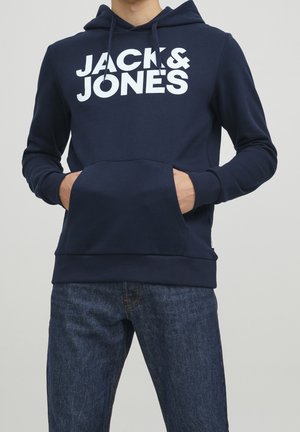 Homme portant un sweat à capuche bleu marine avec le texte "JACK & JONES" en blanc, mains dans la poche avant, associé à un jean bleu foncé.