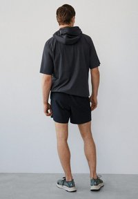 Top negro con capucha de manga corta, con un corte holgado, combinado con pantalones cortos oscuros y zapatillas deportivas grises. Diseño simple, textura suave.