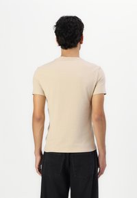 Homme aux cheveux courts et bouclés de dos, portant un t-shirt beige à manches courtes et un jean noir sur fond blanc uni.