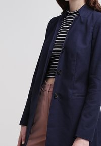 Abrigo azul marino con un diseño estructurado, que presenta un cuello alzado, dos botones y una tela texturizada. Llevado sobre una blusa a rayas.