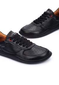 Zapatillas de cuero negro con puntera perforada, cordones negros y acentos rojos. Presentan una suela de goma texturizada y un detalle de costura visible.