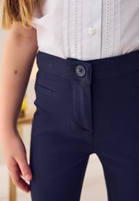 Tumši zili bikses ar gludu tekstūru, ar redzamu pogu aizdari un divām priekšējām kabatām. Valdātas ar baltu kreklu.