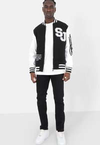 Zwart-wit varsity jack met "SJ" logo, witte mouwen, gestreepte manchetten en geborduurde details. Draagt een wit t-shirt en zwarte jeans.