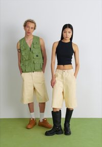 Due modelli si trovano su un pavimento verde. Uno indossa un gilet tattico verde, pantaloni corti gialli e scarpe marroni. L'altro indossa un top nero, pantaloni corti gialli e stivali neri fino al ginocchio.