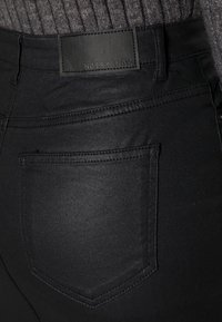 Černé džíny s hladkou texturou, klasickým designem s pěti kapsami a obdélníkovou koženou nášivkou označenou "NOISY MAY" vzadu.