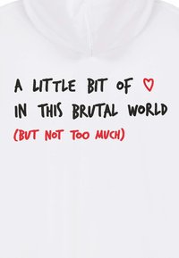 Il testo su un tessuto bianco recita: "Un po' di [simbolo cuore] in questo mondo brutale (ma non troppo)" in lettere nere e rosse.