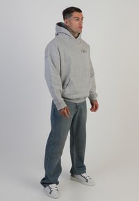 Hanorac gri cu buzunar tip cangur, croială lejeră și logo pe piept, asociat cu blugi albaștri deschiși și adidași albi.