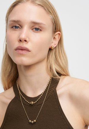 Blonde woman with blue eyes wearing a dark brown sleeveless top and layered gold bead necklaces and matching gold stud earrings.