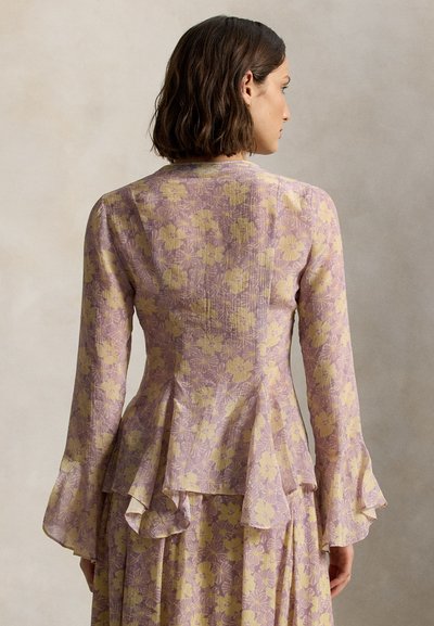 Frau mit kurzen braunen Haaren trägt eine langärmlige Fliederbluse mit gelbem Blumenmuster und ausgestellten Manschetten, steht vor einem neutralen Hintergrund.