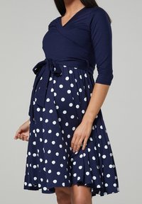Mörkblå klänning med V-ringning och trekvartsärmar, med vita prickar och ett knytbälte i midjan. Utsvängd kjol.