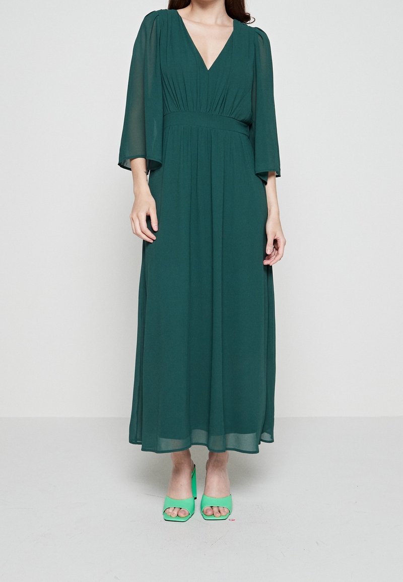 Femme portant une longue robe vert foncé avec des manches 3/4 transparentes et des sandales à talons ouvertes vert clair, debout sur un fond uni.