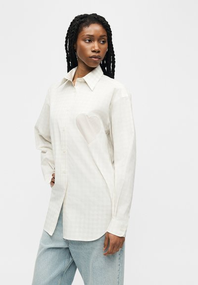 Γυναίκα με μακριά κοτσίδια που φοράει ανοιχτό, άσπρο πουκάμισο με κουμπιά και ένα διακριτικό φιόγκο στην αριστερή πλευρά, και ανοιχτόχρωμο μπλε τζιν παντελόνι.