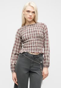 Νεαρή γυναίκα με μακριά ξανθά μαλλιά φοράει ένα καρό τοπ με λάστιχο και σκούρο γκρι τζιν με διακοσμητική ασημένια αλυσίδα ζώνης σε απλό φόντο.