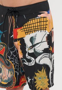 Lautashorts dengan latar belakang hitam menampilkan desain besar yang terinspirasi tato dengan motif ular, burung, jaring, dan matahari yang berwarna-warni.