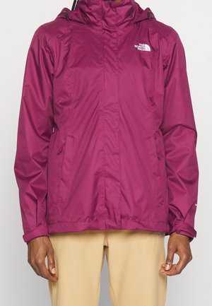 Personne portant une veste à capuche magenta North Face avec des poches zippées et un pantalon beige, debout devant un fond uni.