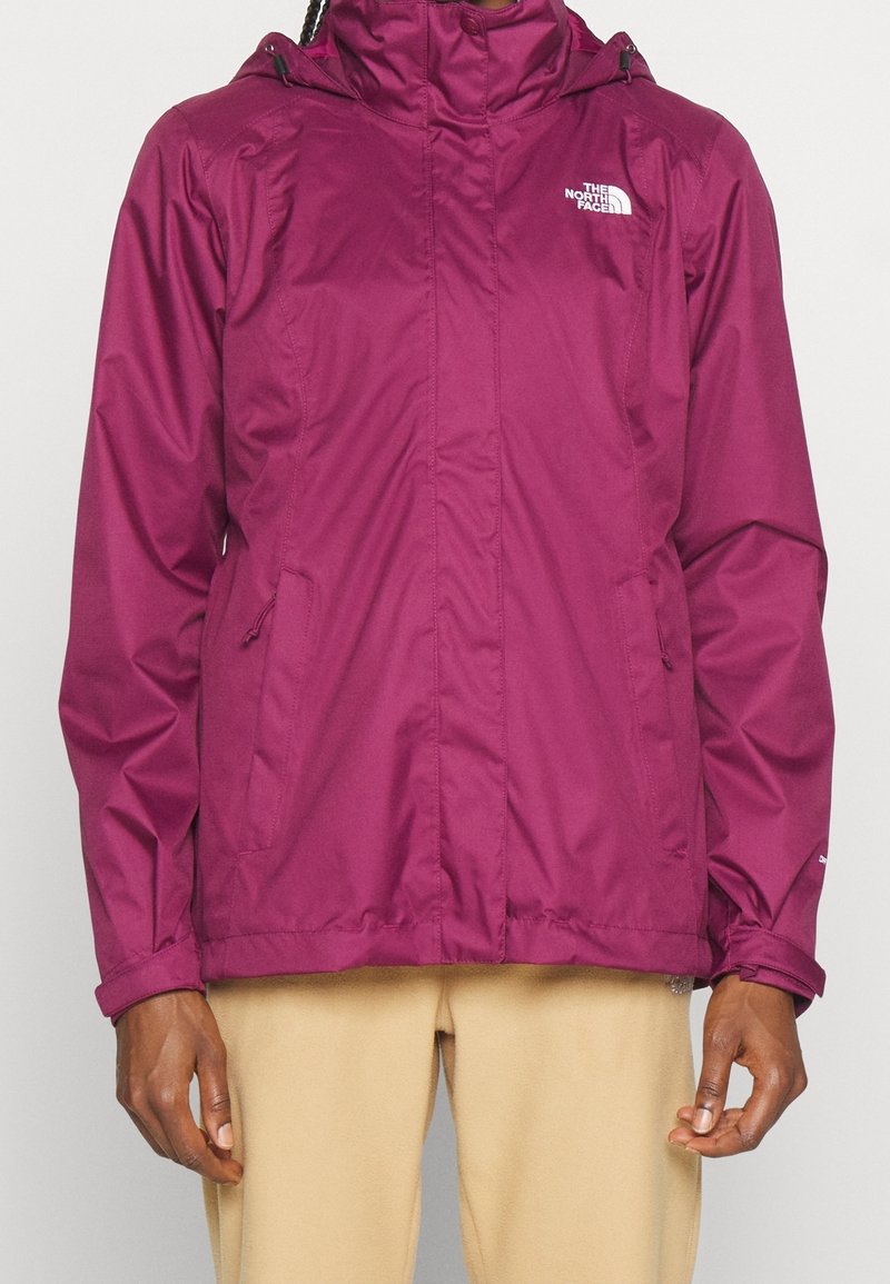 Personne portant une veste à capuche magenta North Face avec des poches zippées et un pantalon beige, debout devant un fond uni.