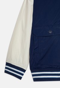 Veste marine et crème avec des poignets élastiqués à rayures bleues, une poche sur le côté et une texture de tissu lisse et léger.