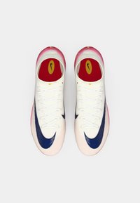 Witte sportschoenen met een marineblauwe swoosh, een vervaagde roze neuskap en een rode binnenzool met een gele logo. Gestructureerde bovenkant en veters aanwezig.