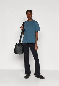 Kvinna som står vänd mot kameran iförd en teal t-shirt, svarta byxor, svarta sneakers och bär en svart axelväska mot en vit bakgrund.