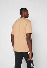 T-shirt en coton beige à manches courtes et à coupe décontractée, porté avec un pantalon noir comportant des rayures blanches sur les côtés, vu de dos.