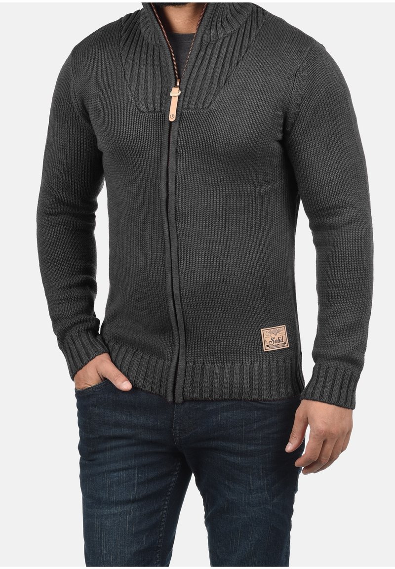 Homme portant un pull en tricot gris foncé avec fermeture éclair, col et poignets côtelés, associé à un jean bleu foncé, une main dans la poche.