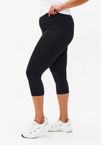 Black capri leggings made of stretch fabric, fitted design, smooth texture, with ankle-length cut. Paired with white athletic sneakers.