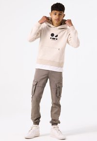 Felpa beige con cappuccio e grafica nera, tasca frontale, abbinata a pantaloni cargo grigi chiari con più tasche e polsini elastici. Sneakers bianche.