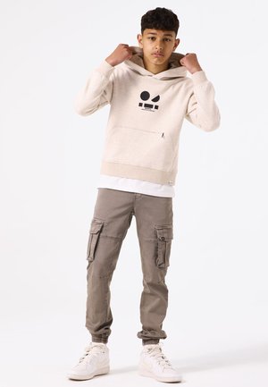 Felpa beige con cappuccio e grafica nera, tasca frontale, abbinata a pantaloni cargo grigi chiari con più tasche e polsini elastici. Sneakers bianche.