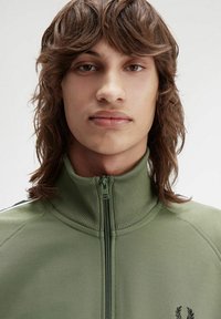 Giacca zip-up in tessuto verde oliva uniforme, con colletto alto, texture a coste e logo ricamato sul lato sinistro.