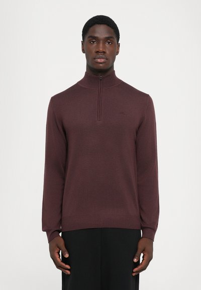 Jeune homme portant un pull à col zippé bordeaux et un pantalon noir, se tenant contre un fond blanc uni, tourné vers l'avant.