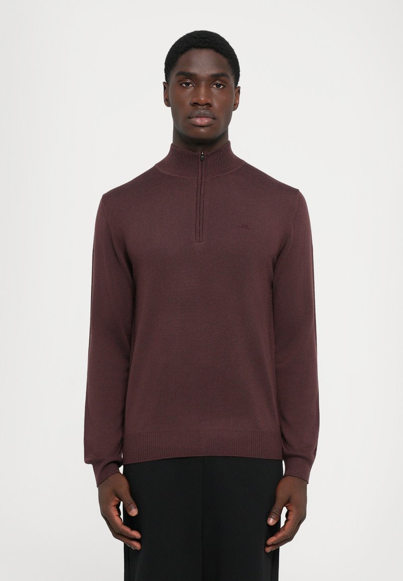 Jeune homme portant un pull à col zippé bordeaux et un pantalon noir, se tenant contre un fond blanc uni, tourné vers l'avant.