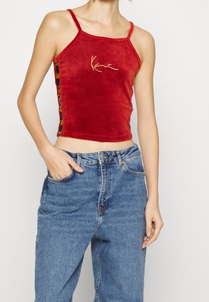 Top corto in velluto rosso con spalline sottili, dotato di un logo dorato e accenti laterali colorati e a fantasia. Abbinato a jeans blu a vita alta.
