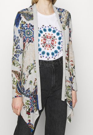 Vrouw die een grijze cardigan met bloemenprint draagt over een wit T-shirt met een kleurrijk cirkelontwerp en de tekst "De toekomst is nu", gecombineerd met een hoge taille zwarte jeans.