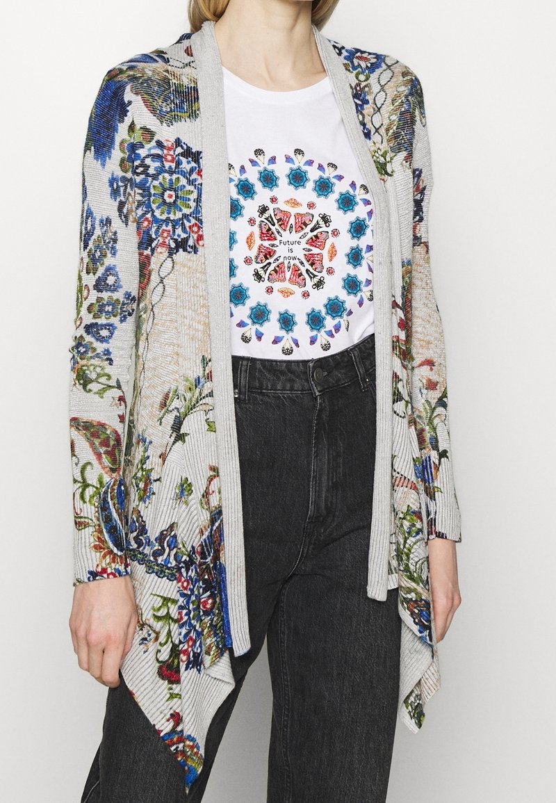 Femme portant un cardigan gris à fleurs sur un t-shirt blanc avec un motif circulaire coloré et le texte « L'avenir, c'est maintenant », assorti à un jean noir taille haute.