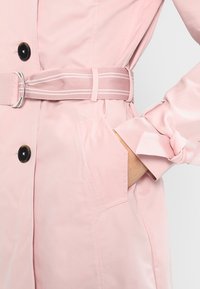 Różowy płaszcz trencz z paskiem w talii, zapięciem na guziki i mankietami ozdobionymi kokardkami. Gładka tkanina o satynowej fakturze.