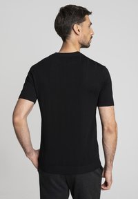 Mężczyzna z krótkimi ciemnymi włosami w dopasowanej czarnej koszulce z krótkim rękawem i ciemnych spodniach, stoi z jedną ręką na biodrze, z twarzą odwróconą w stronę.