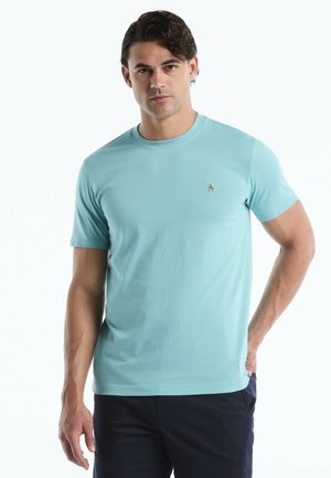 Man staat recht vooruit met een hand in de zak, draagt een lichtblauw kort mouw T-shirt en donkere broek.