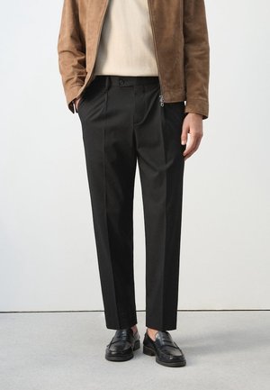 Mann trägt schwarze maßgeschneiderte Hose, schwarze Slipper, beigen Pullover und braune Wildlederjacke vor einfarbigem Hintergrund.