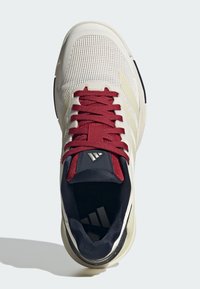 Scarpa da ginnastica beige con tomaia in mesh, interno blu scuro, lacci color granata e tre strisce bianche sul lato.