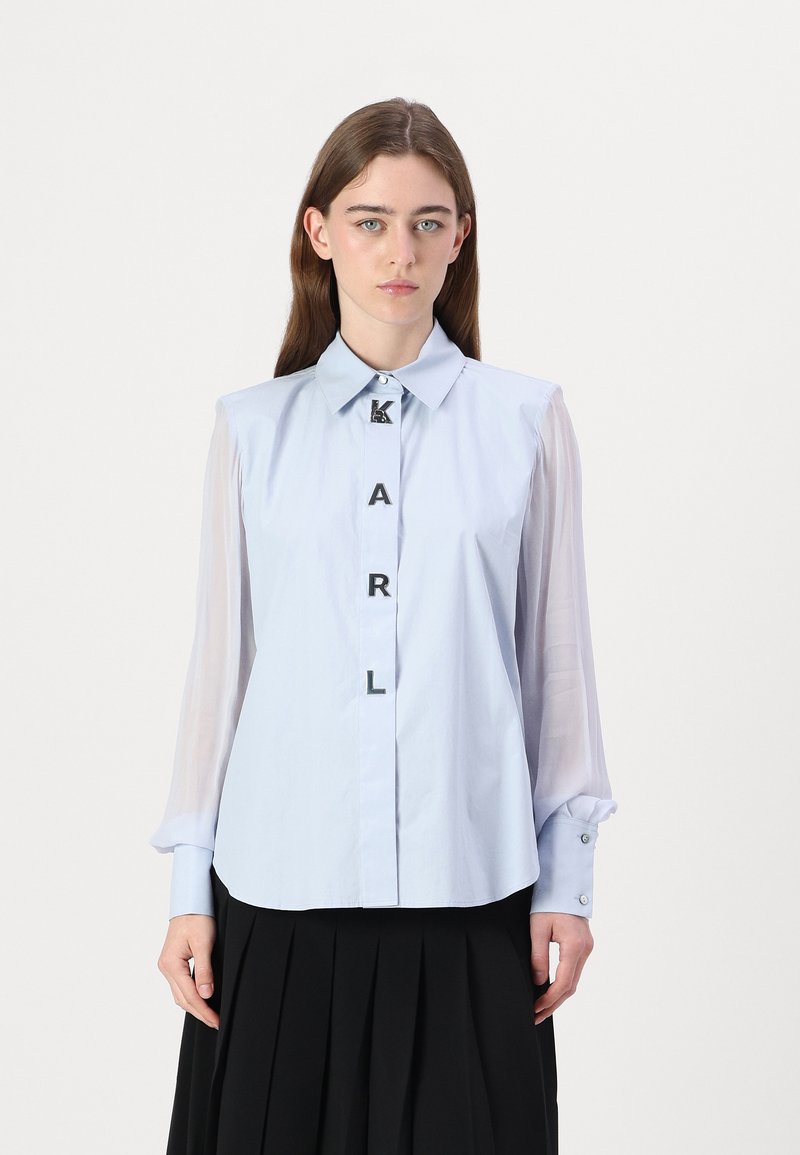 Camicia button-up azzurra chiara con maniche trasparenti, caratterizzata da accenti neri a forma di lettere che formano la parola "KARL" sulla parte frontale. Completata con un colletto classico.
