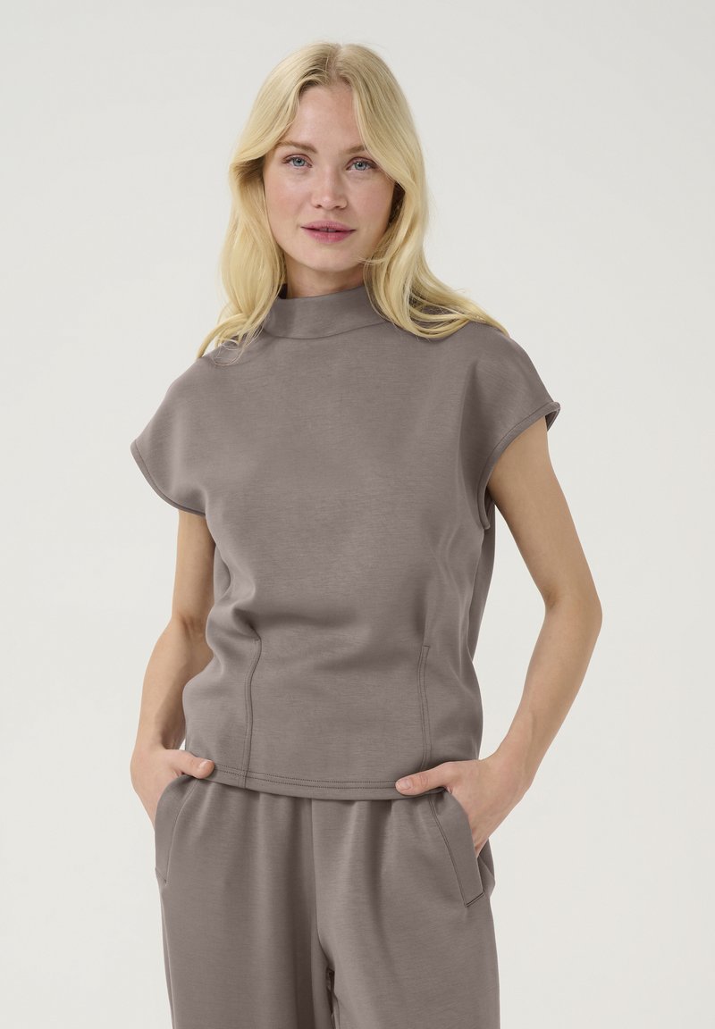 Top grigio a maniche corte con colletto alto, realizzato in tessuto morbido. Presenta dettagli sulle cuciture e tasche laterali. Indossato con pantaloni grigi coordinati.