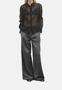 Mustat läpikuultavat napitettavat paidat, jotka on kerrostettu tummanvärisen topin päälle, yhdistettynä korkeavyötäröisiin, leveälahkeisiin harmaisiin housuihin, jotka on valmistettu teksturoidusta kankaasta.