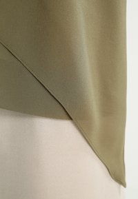 Olivgrön topp med en mjuk textur, med en diagonal söm som övergår till en ljusbeige panel längst ner.