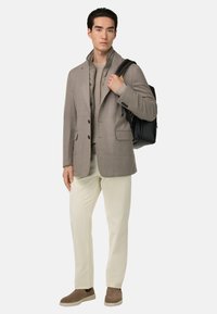 Blazer beige con bottoni scuri, maglione lavorato crema, pantaloni beige chiaro e scarpe marroni. Zaino nero sospeso su una spalla.